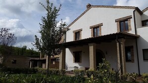 A two-story house with a covered porch, brickwork, and a garden with various plants. - Casali Costadoro (Acquaviva Picena)