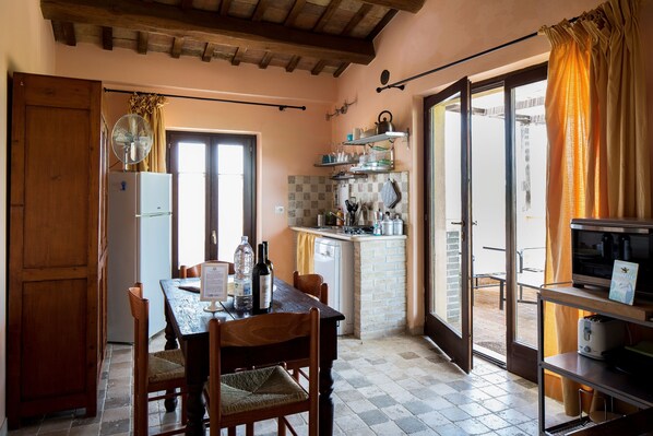 A kitchen with a dining table, chairs, a refrigerator, a sink, and a window with curtains. - Casali Costadoro (Acquaviva Picena)