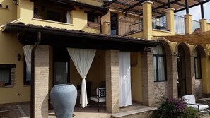 Un edificio de dos pisos con un patio cubierto, que incluye un jarrón grande y una silla.