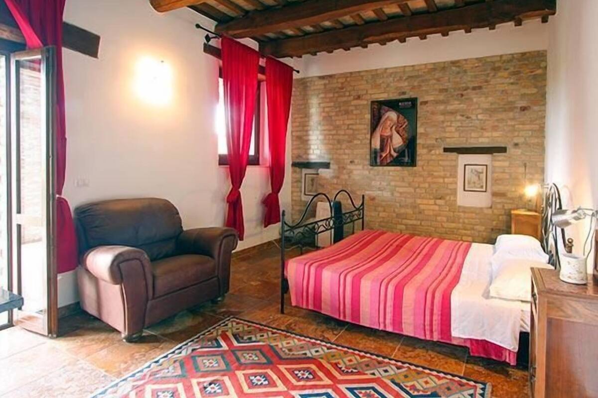 A bedroom with a bed, a chair, a stone wall, and a window with red curtains.