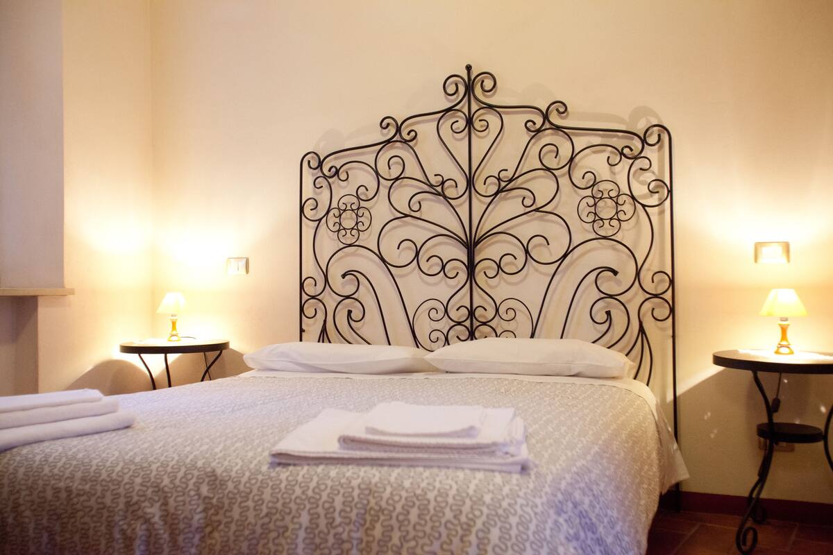 Una cama bien hecha con una colcha estampada, dos mesitas de noche con lámparas y un cabecero decorativo de metal.