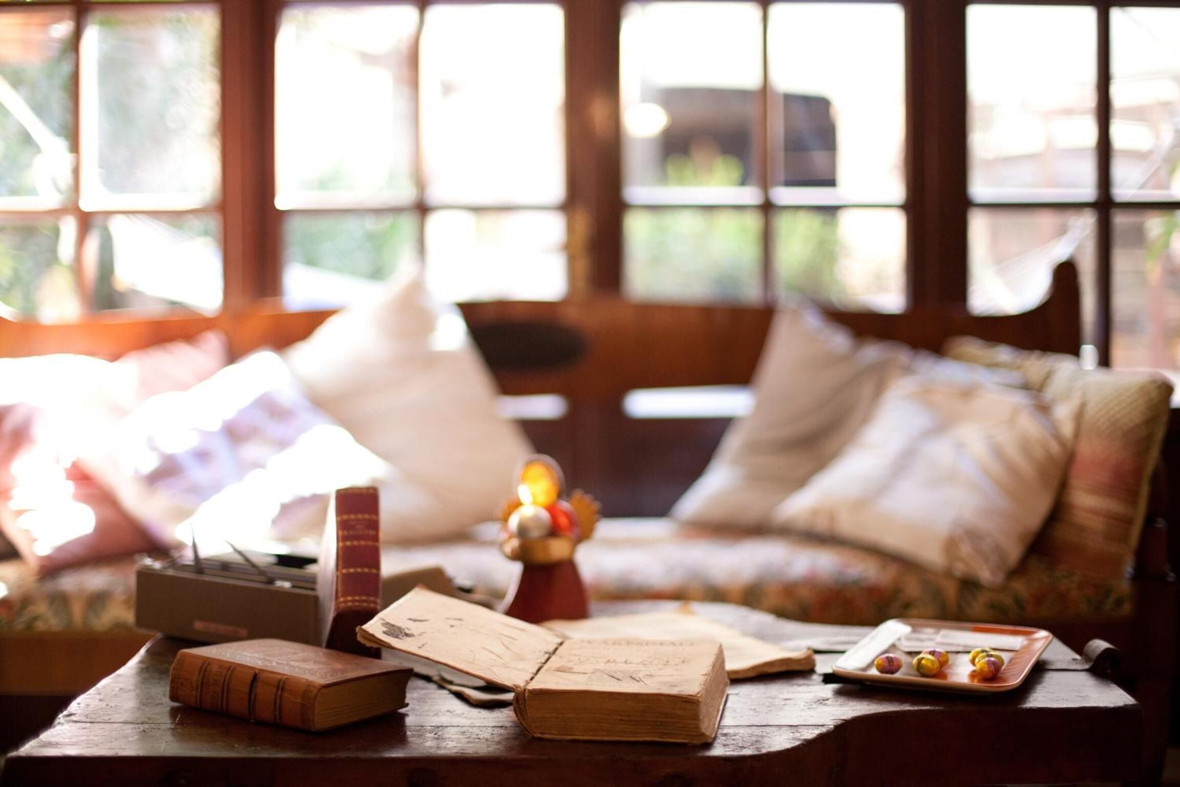 A cozy room with a wooden table, books, a lamp, and a window with a view of greenery.