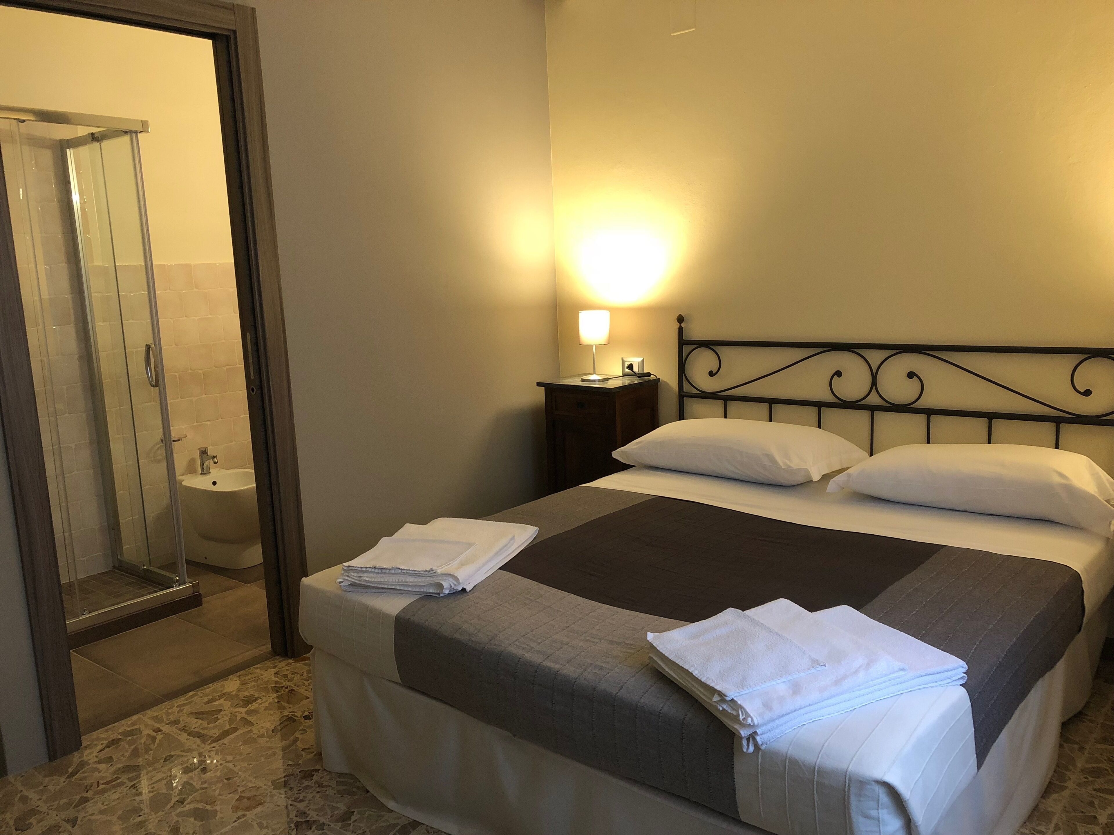 A hotel room with a bed, a bedside table with a lamp, a bathroom with a glass shower enclosure, and a toilet.