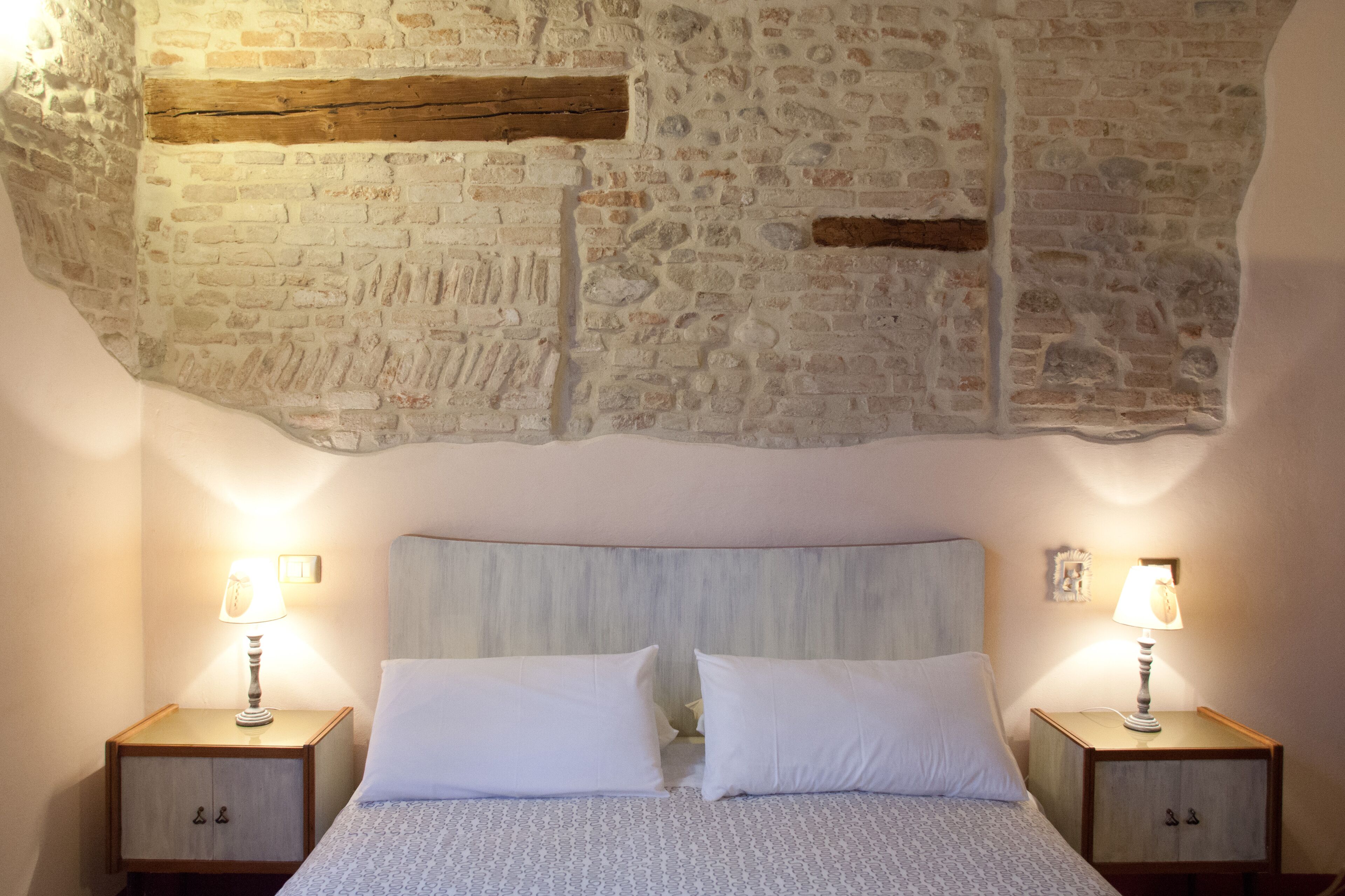 Une chambre avec un mur en pierre, un lit avec une literie blanche, deux tables de chevet avec des lampes et une tête de lit en bois.