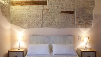 Une chambre avec un mur en pierre, un lit avec une literie blanche, deux tables de chevet avec des lampes et une tête de lit en bois.