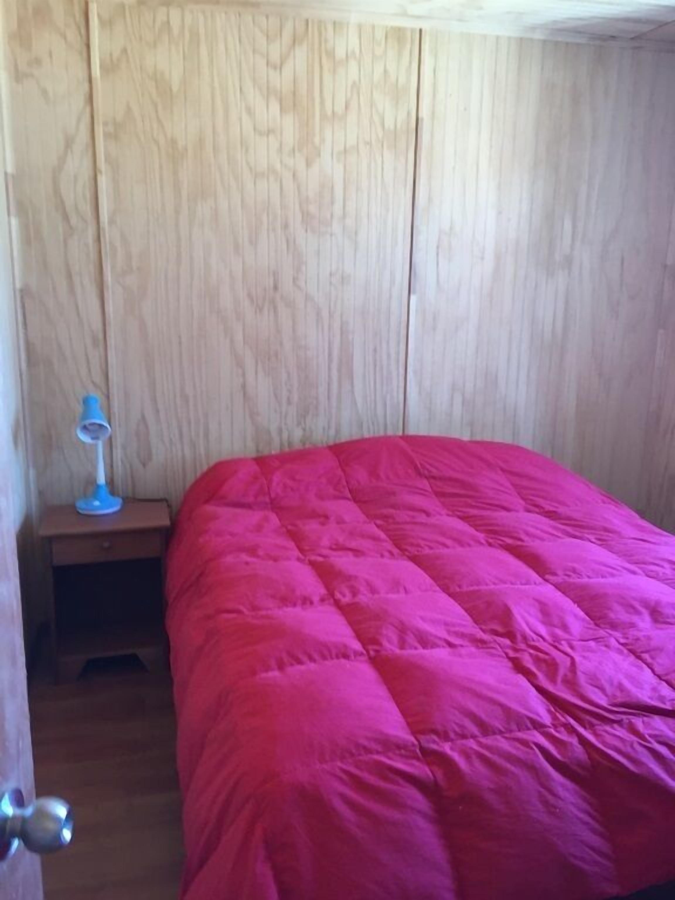 A room with a wooden paneled wall, a red bedspread, a small bedside table with a lamp, and a door with a round handle.