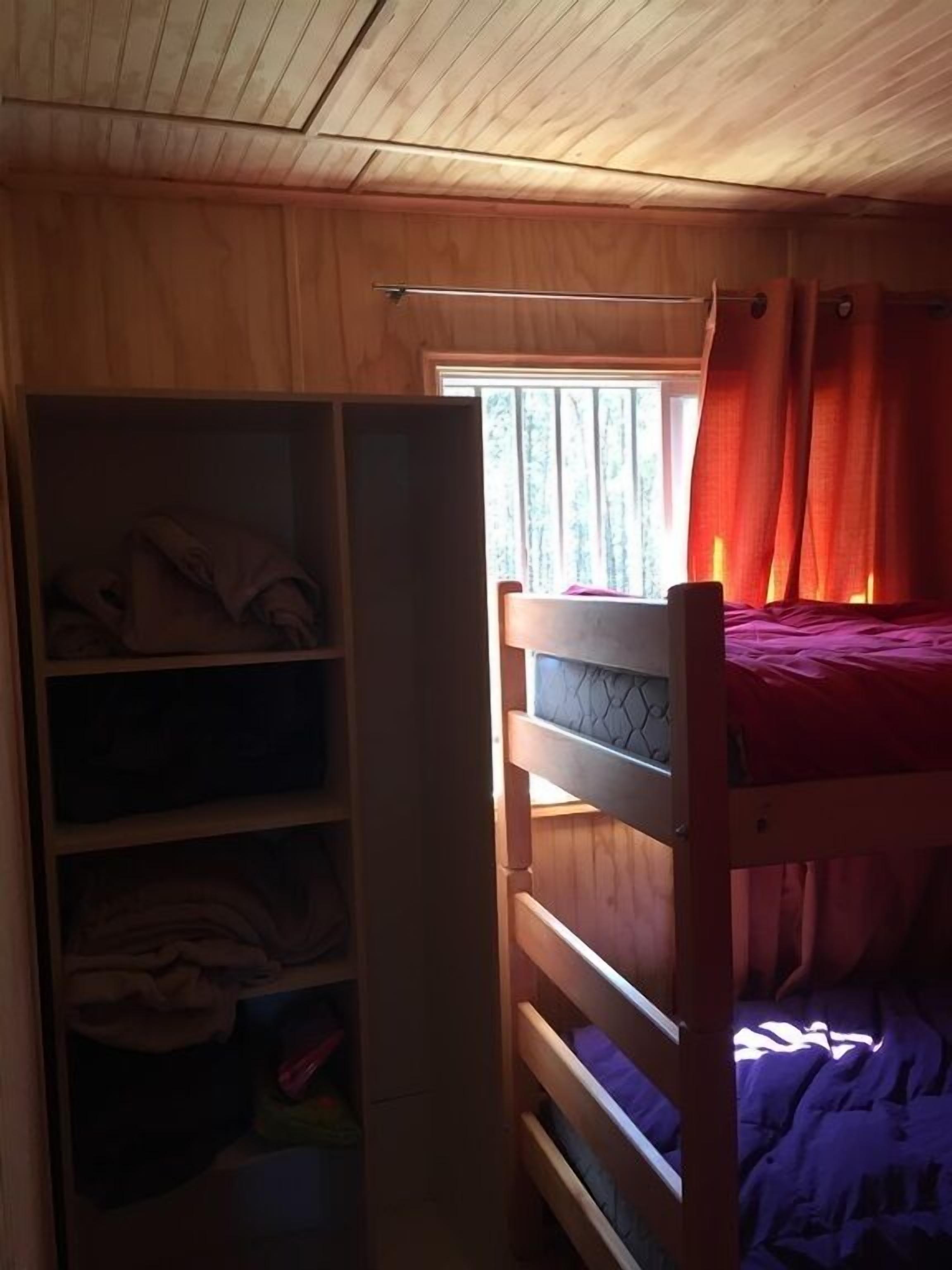 Una cama literera con colchón rojo, un armario de madera con estantes y una ventana con cortinas.