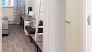 A modern, compact room with a wooden floor, a bed with a black and white patterned pillow, a desk with a lamp, and a wardrobe with hanging space. - Innotelli Apartments (Helsinki)