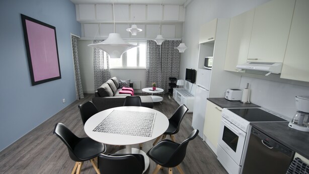 A modern kitchen with white appliances, a dining area with a round table and black chairs, and a living area with a sofa and curtains. - Innotelli Apartments (Helsinki)