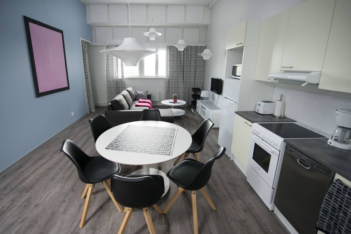 A modern kitchen with white appliances, a dining area with a round table and black chairs, and a living area with a sofa and curtains.