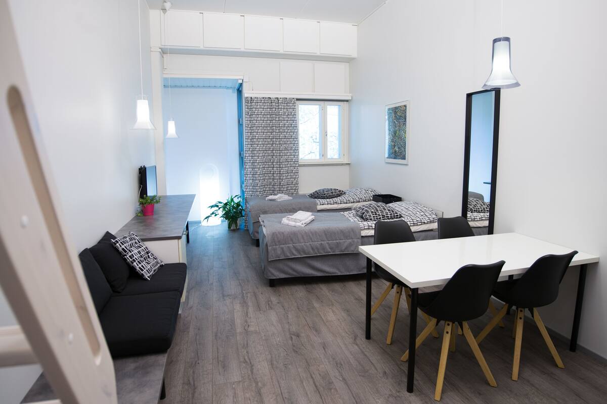 A modern, well-lit room with a bed, a dining table with chairs, a small kitchenette, and a large mirror.