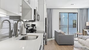A modern kitchen with a sink, stove, and refrigerator. There is a gray sofa and a small table with a lamp. A large window with curtains is visible in the background. - Sentral East Austin at 1630 E. Sixth (Austin)