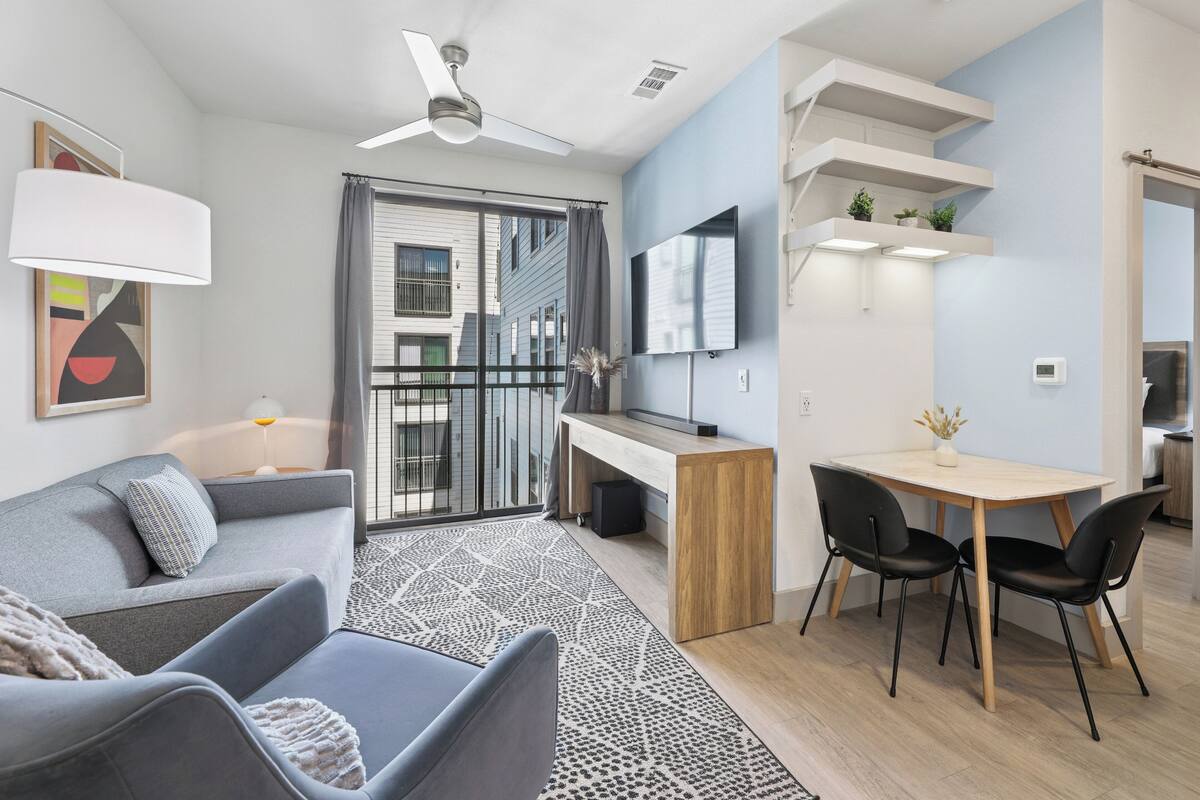 Un salon moderne avec un canapé gris, une table à manger avec deux chaises, un téléviseur fixé au mur et un ventilateur de plafond.