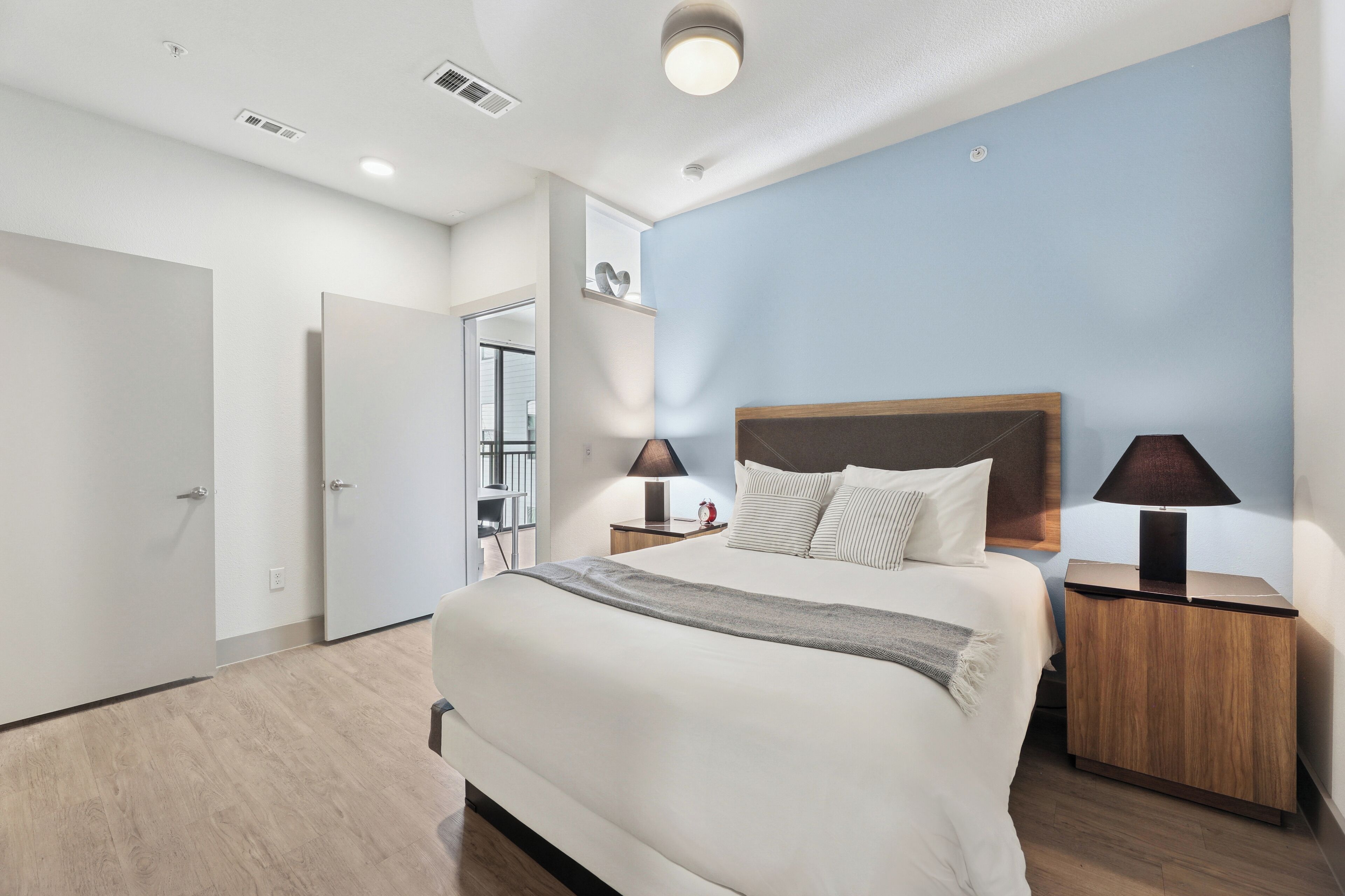 A modern bedroom with a large bed, two bedside tables with lamps, a wooden headboard, and a light blue accent wall.