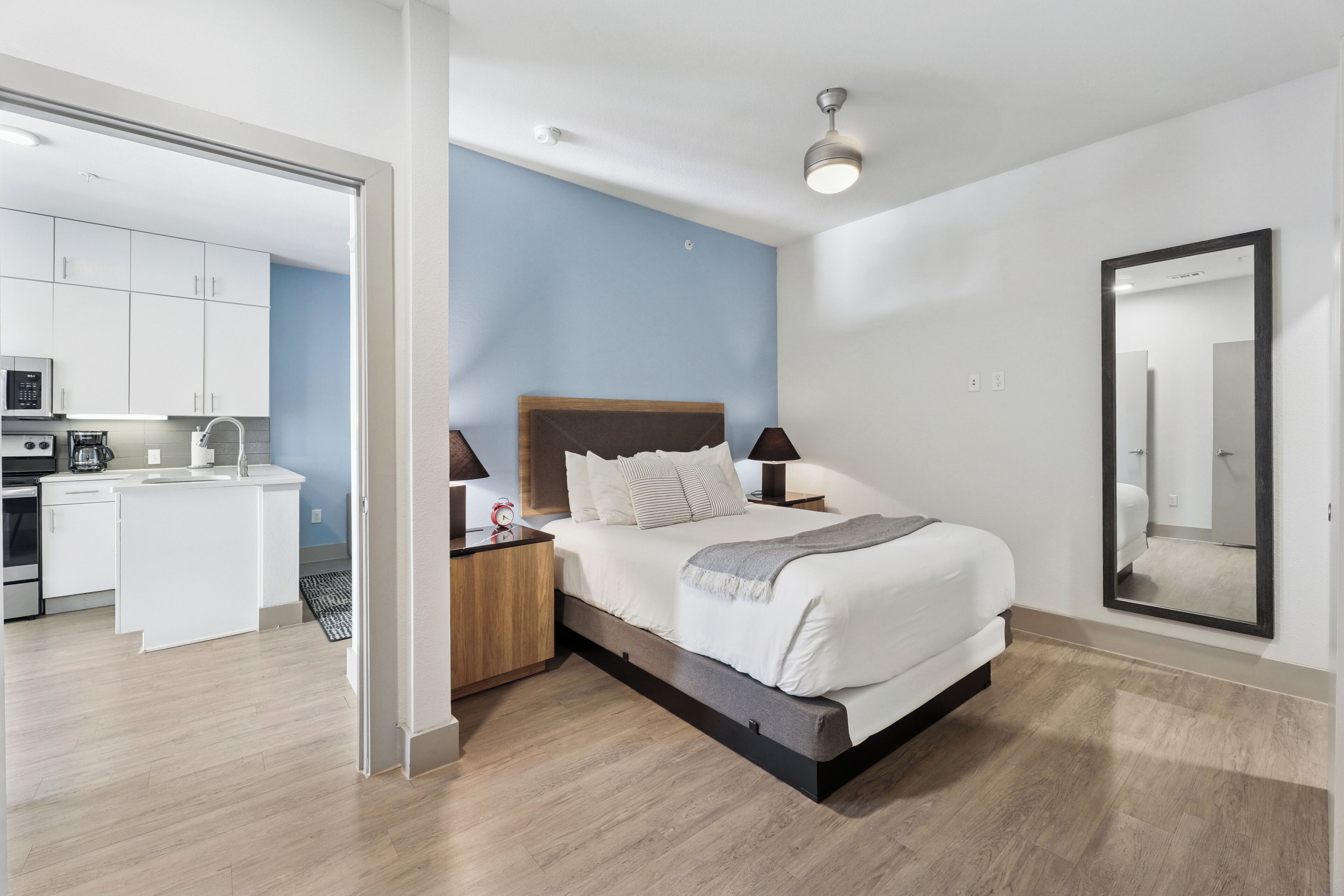 A modern bedroom with a bed, bedside tables, a nightstand, a large mirror, and a view into the adjacent kitchen area.