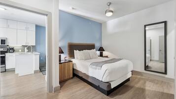 Un dormitorio moderno con cama, mesitas de noche, un espejo grande y vistas al área de la cocina contigua.