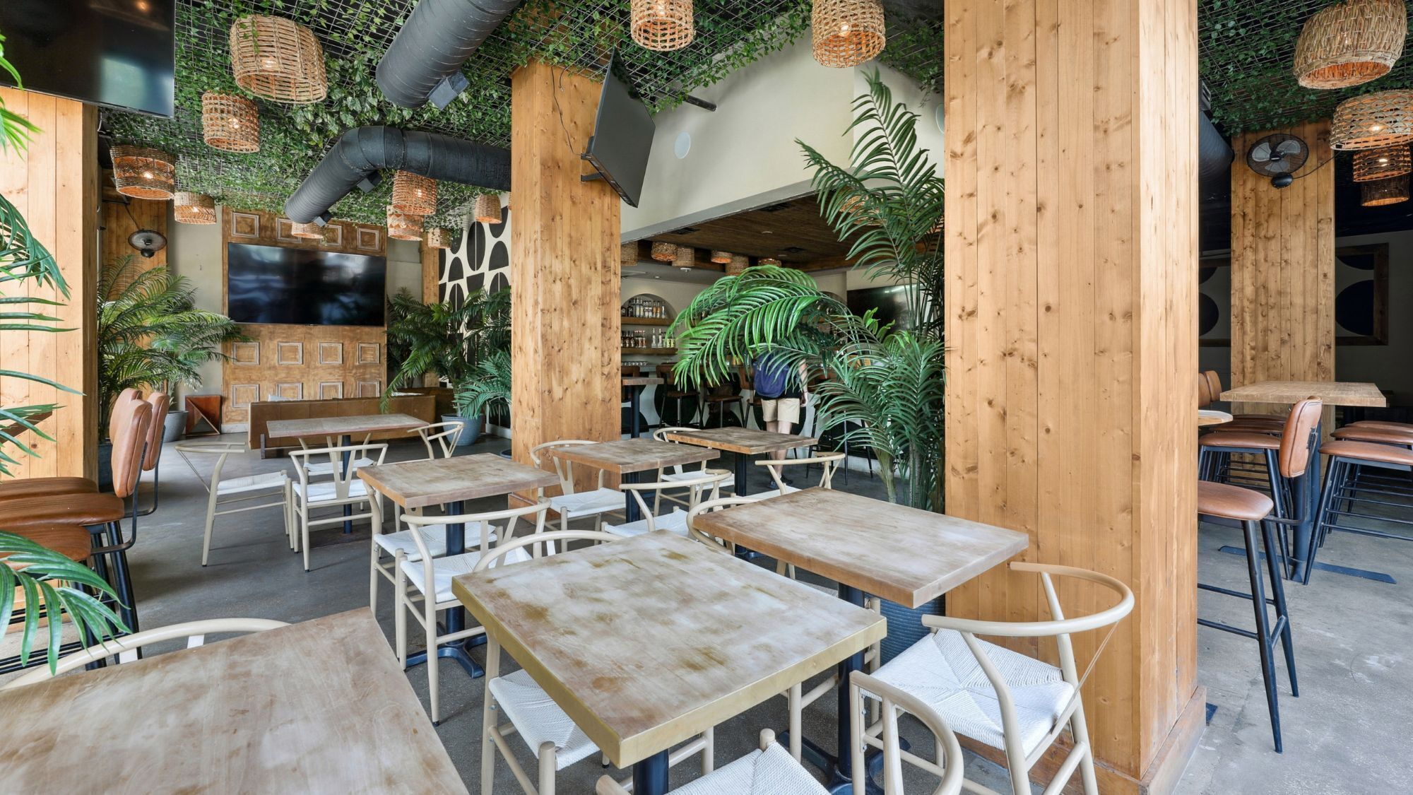 A restaurant interior with wooden tables and chairs, a TV screen, and various plants.