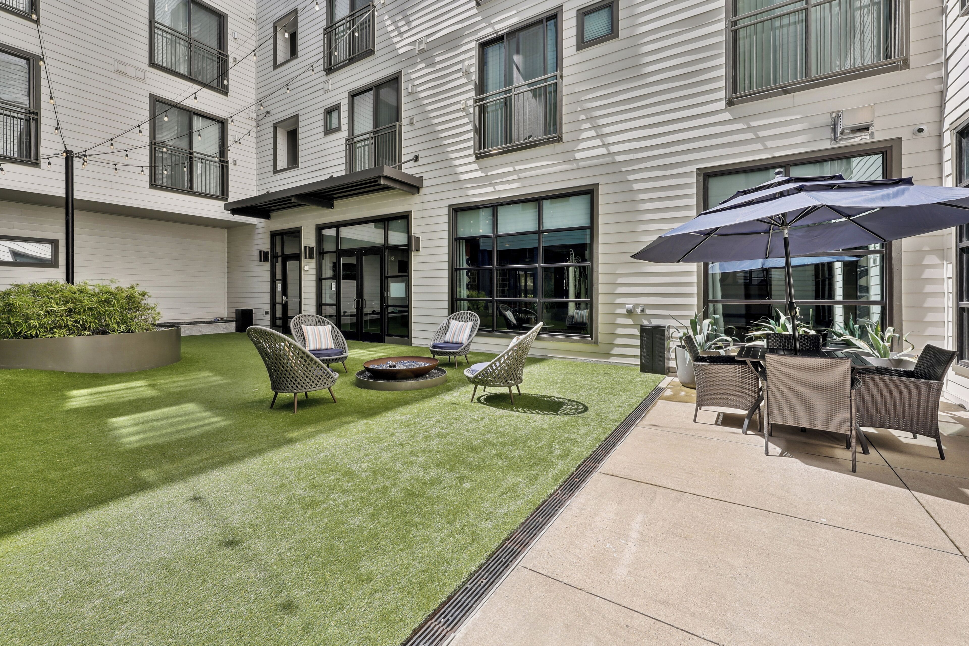 Une terrasse extérieure moderne, dotée d’un gazon synthétique, de meubles en osier et d’un grand parasol.