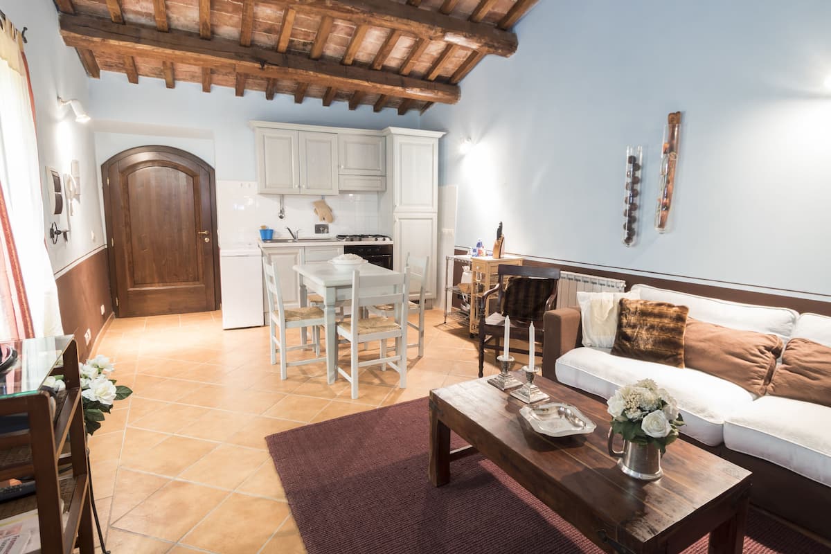 Una sala de estar acogedora con una mesa de centro de madera, sofá blanco y una zona de cocina con armarios blancos y una puerta de madera.