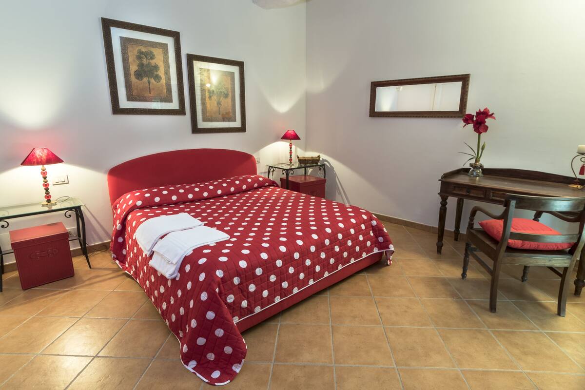 Un dormitorio con cama, escritorio, silla y una mesita con un jarrón de flores.
