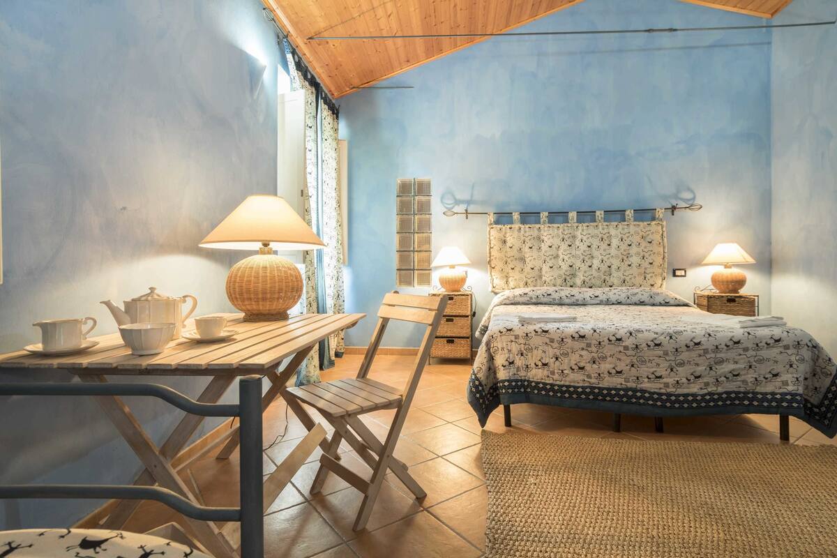 Un dormitorio acogedor con cama, lámparas de noche, una mesita de madera con juego de té y una silla.
