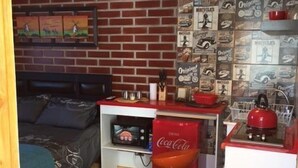 Ein gemütlicher Raum mit einer Wand im Backsteinmuster, einem Bett mit Kopfteil, einem Schreibtisch mit einem roten Coca-Cola-Kühler, einer gelben Pendelleuchte und einem roten Wasserkocher auf einer weißen Arbeitsplatte.