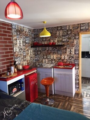 Una cocina compacta con pared de ladrillo, un refrigerador rojo, una lámpara colgante amarilla y un revestimiento de pared estampado.