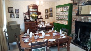 Ein Esszimmer mit Holztisch, gedeckt für eine Mahlzeit, umgeben von Stühlen. Im Hintergrund ist ein grünes Bücherregal zu sehen, und rechts befindet sich ein Kamin.