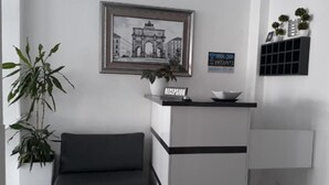A reception area with a framed picture of a historical building, a reception desk, a potted plant, and a sign with a phone number. - Ceko Hotel (Sarandë)