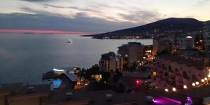 Une vue d’une ville côtière au crépuscule, avec des bâtiments, un port de plaisance et une montagne lointaine.