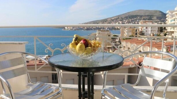 A balcony with a table and chairs, a bowl of fruit, and a view of the sea and buildings. - Ceko Hotel (Sarandë)