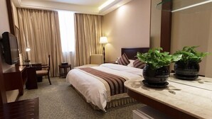 Quarto de hotel com cama, mesa de cabeceira, luminária, televisão, escrivaninha, cadeira e duas plantas em vasos.