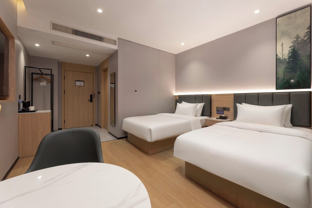 Ein Hotelzimmer mit zwei Betten, einem Schreibtisch und einem Stuhl. An der Wand hängt ein Gemälde und eine Tür führt in einen weiteren Raum.