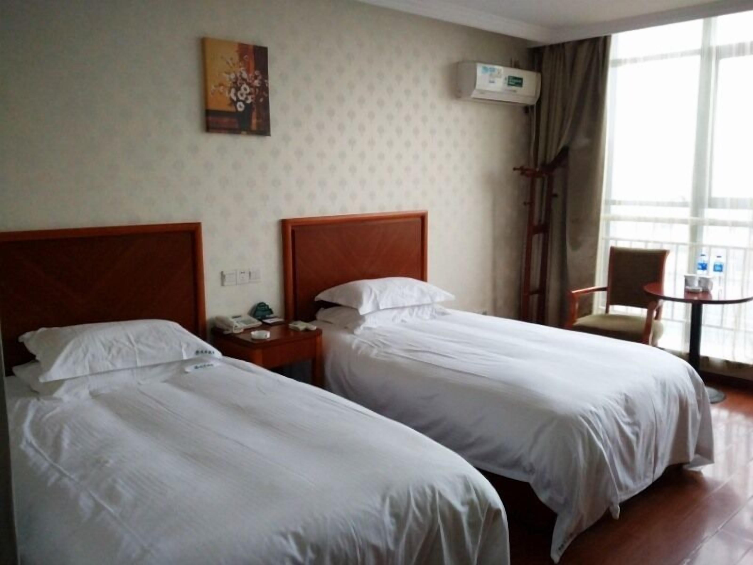 A hotel room with two beds, a small table with a chair, an air conditioning unit, and a window with curtains.