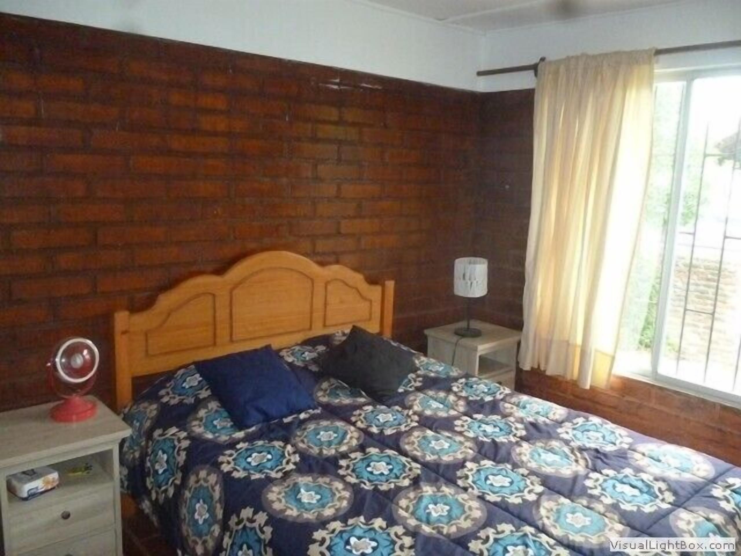 Ein Schlafzimmer mit einer Ziegelwand, einem hölzernen Bettgestell, einer gemusterten Bettdecke, einem Nachttisch mit Lampe und einem Fenster mit Vorhängen.