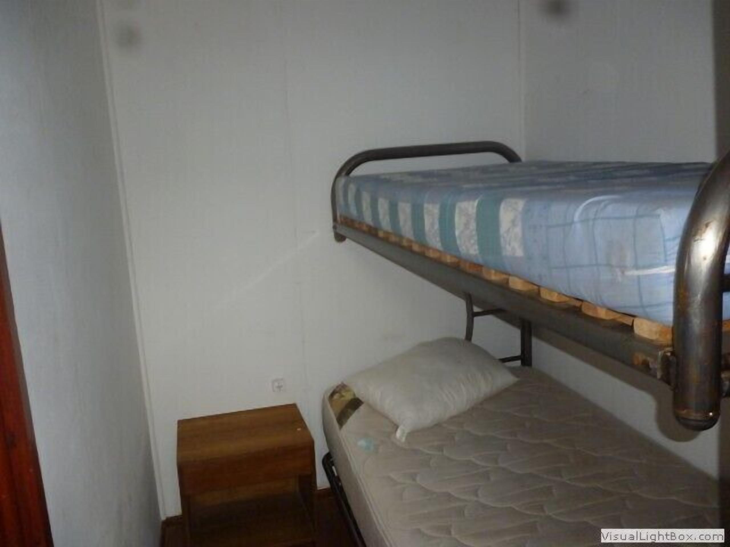 Quarto pequeno e estreito com uma cama de solteiro e uma cama individual. Há um banquinho de madeira e uma pequena mesa.