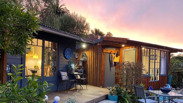 Una cabaña acogedora con fachada de madera, un patio con mobiliario exterior y un jardín con plantas en macetas y una pequeña mesa.