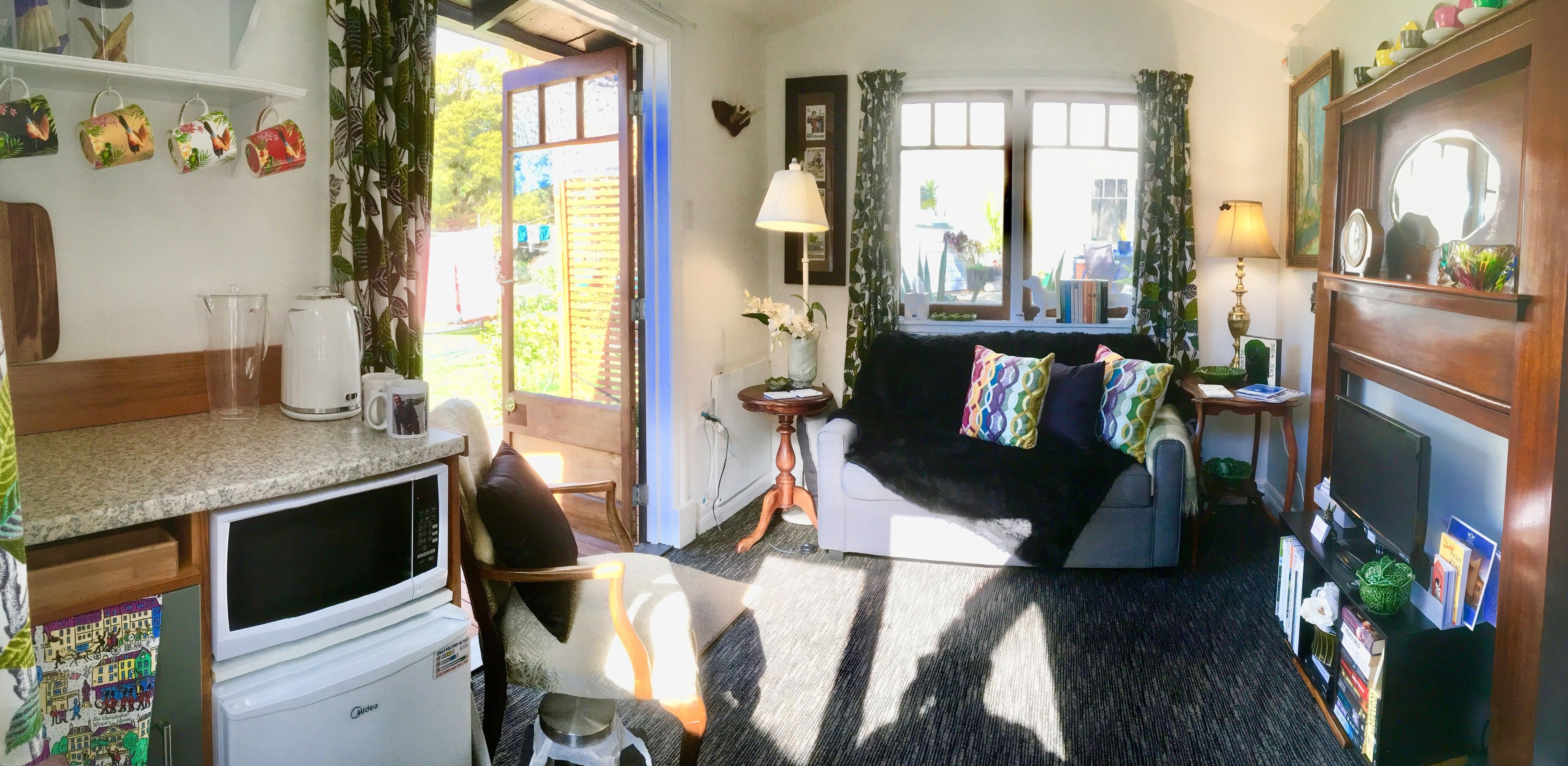 Un espacio habitable compacto con kitchenette, un sofá con cojines coloridos, un televisor y una ventana con cortinas.