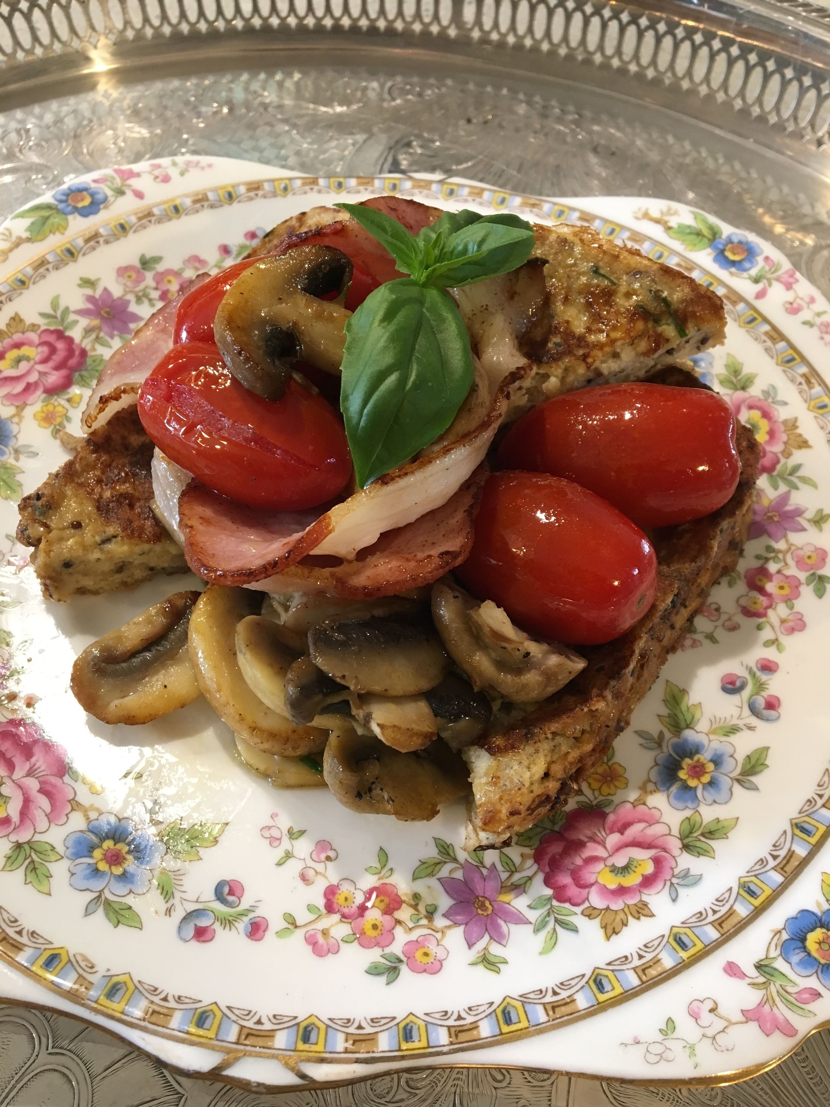 Un plato con un sándwich cubierto de tomates, champiñones y tocino, adornado con una hoja de albahaca.