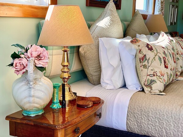 Una cama bien hecha con cubrecama acolchado, una lámpara de noche y un jarrón con flores sobre una mesita de madera.