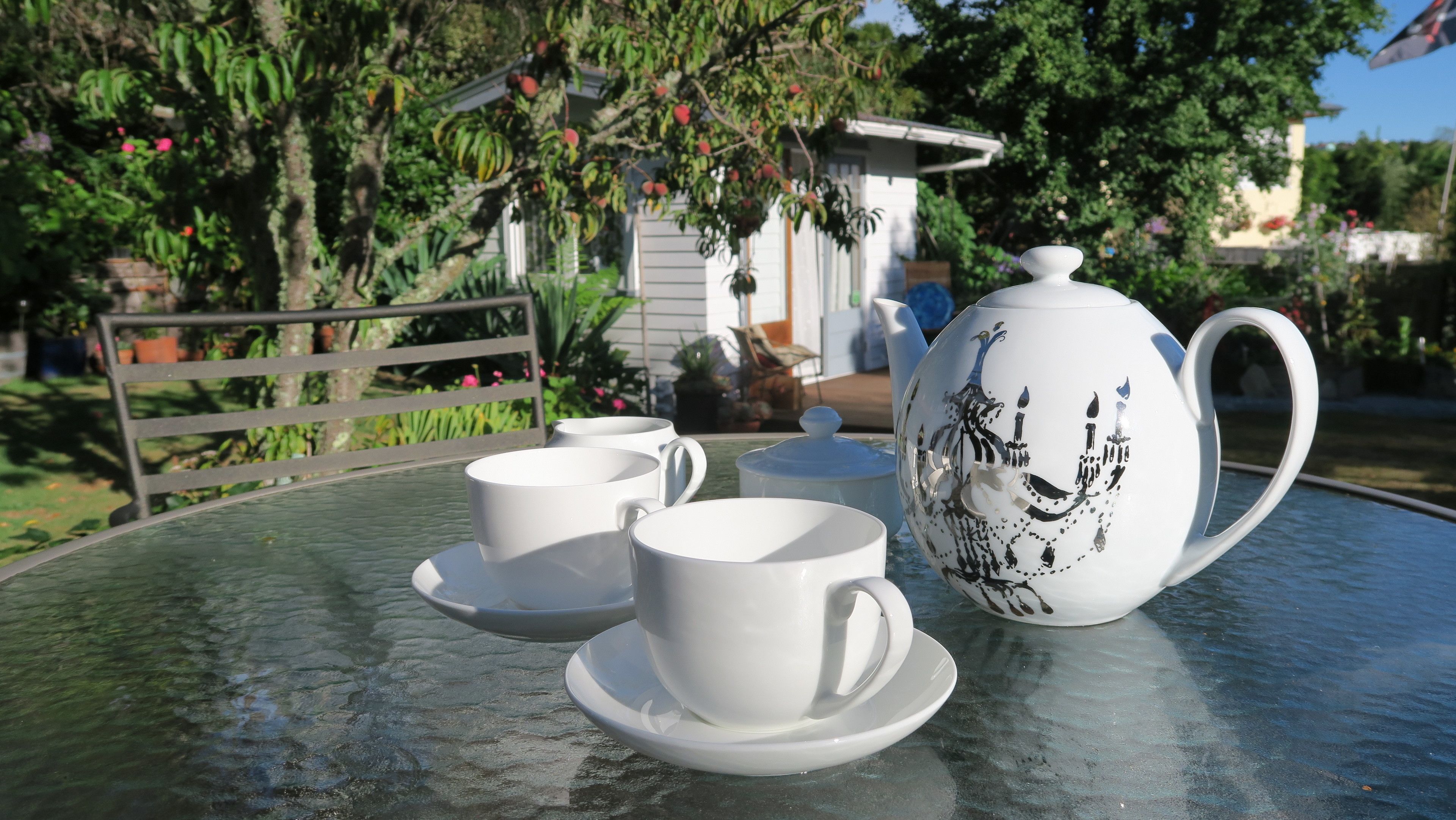 A table with a teapot and two cups on a glass table in a garden setting.