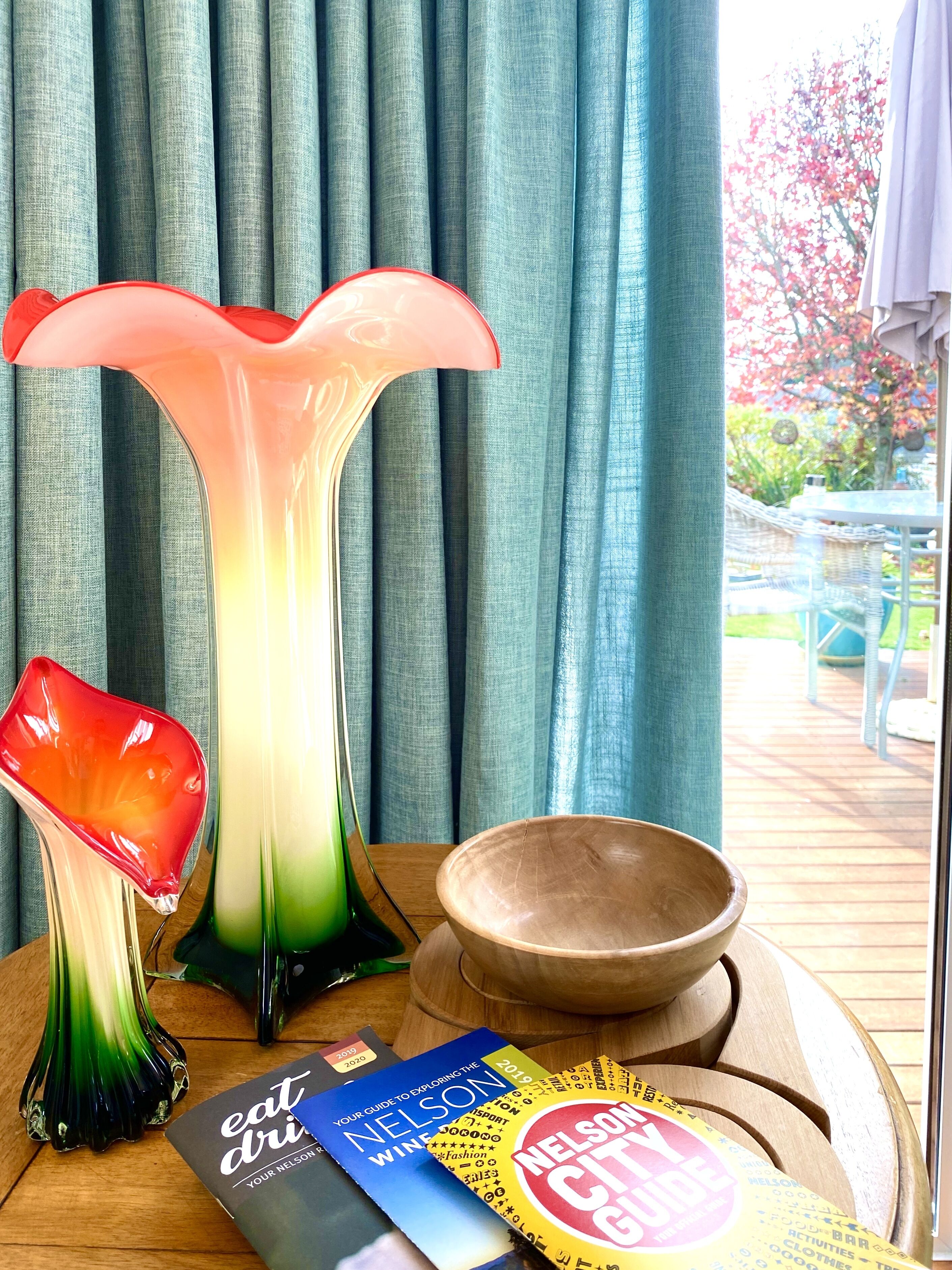 A table with a glass vase, a bowl, and a magazine on a wooden table with a view of a garden through a window.