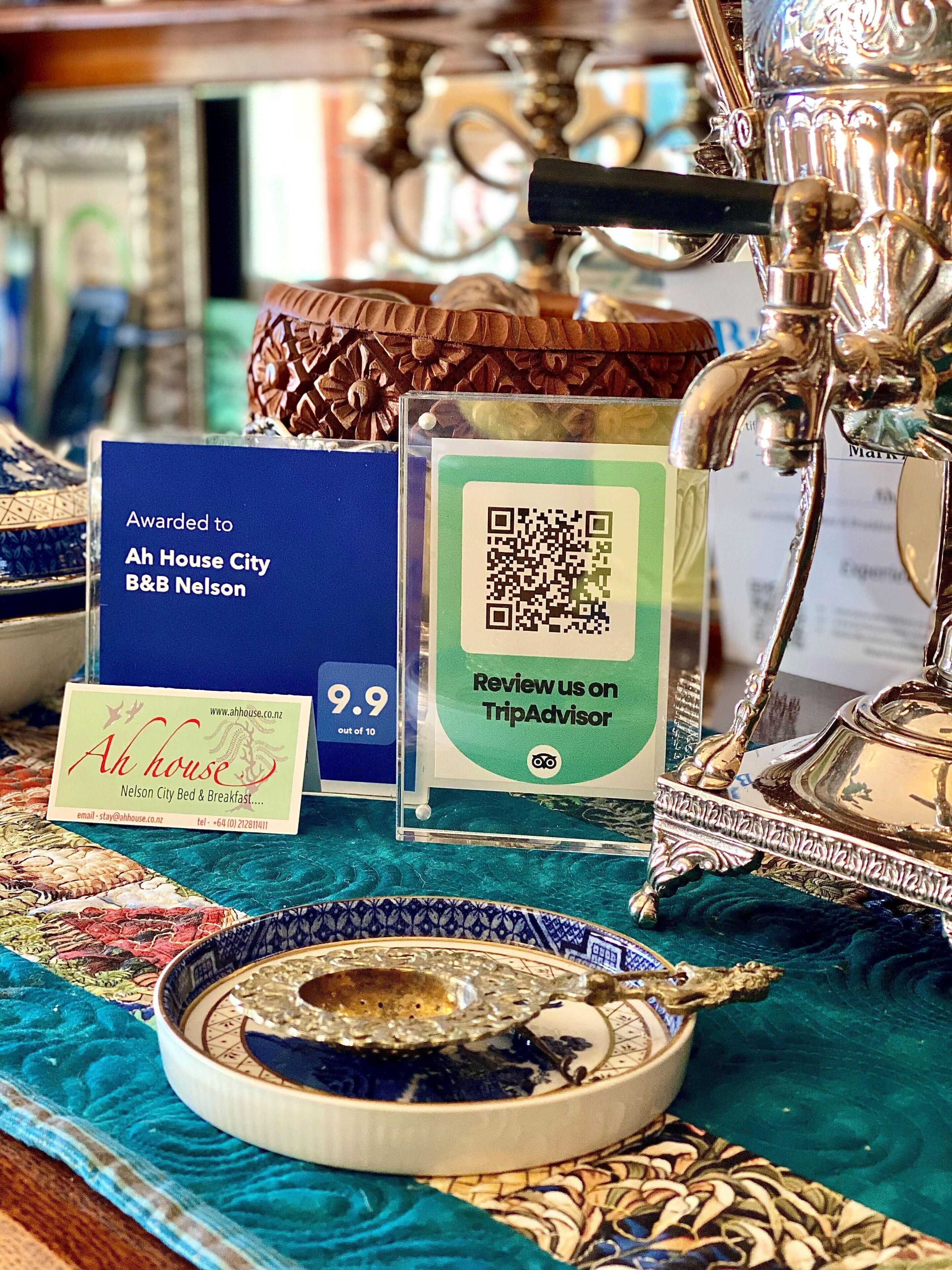 Una mesa con un plato decorativo, un letrero de Ah House City B&B, un código QR de TripAdvisor y una lámpara de latón.