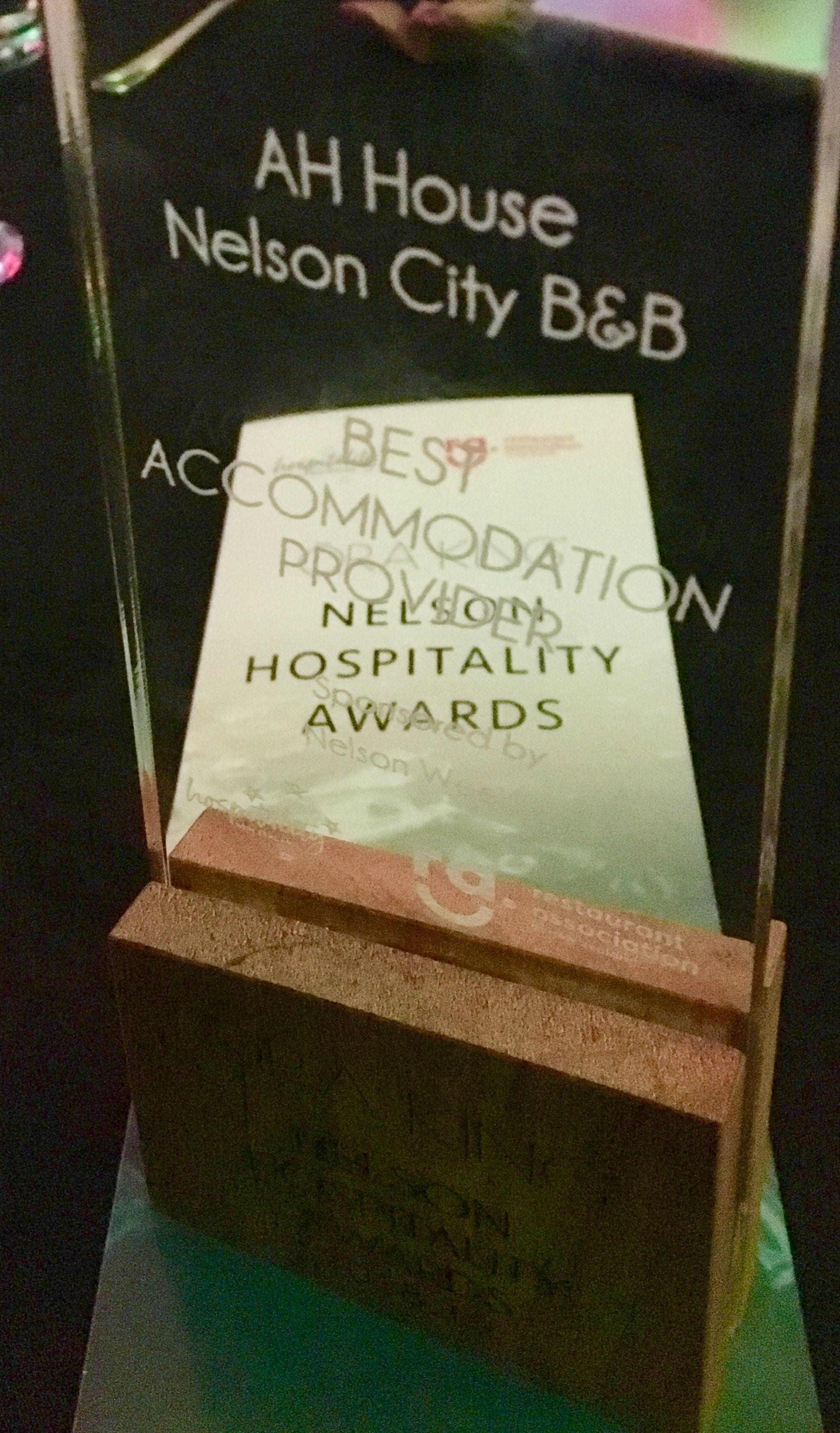 Kehyksiin asetettu palkintalaatta AH House Nelson City B&B -majoitusliikkeelle, jossa korostetaan sen saamista tunnustuksista parhaana majoituspaikkana Nelson Hospitality Awards -palkintogaalassa.