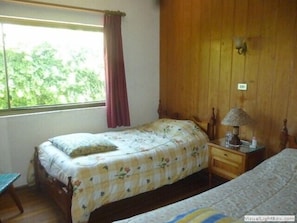 Quarto com cama, mesa de cabeceira, luminária e janela com vista para o verde.