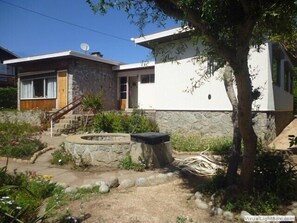 A two-story house with a stone facade and a white upper section. There is a small garden with a stone planter and a tree. - Casa Pescadores Poniente Algarrobo 1030 (Algarrobo)