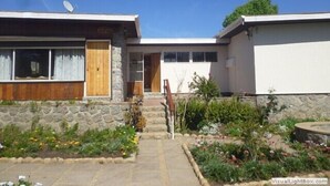 Une maison à deux étages avec une façade en pierre, une antenne parabolique et un jardin où poussent diverses plantes et fleurs.