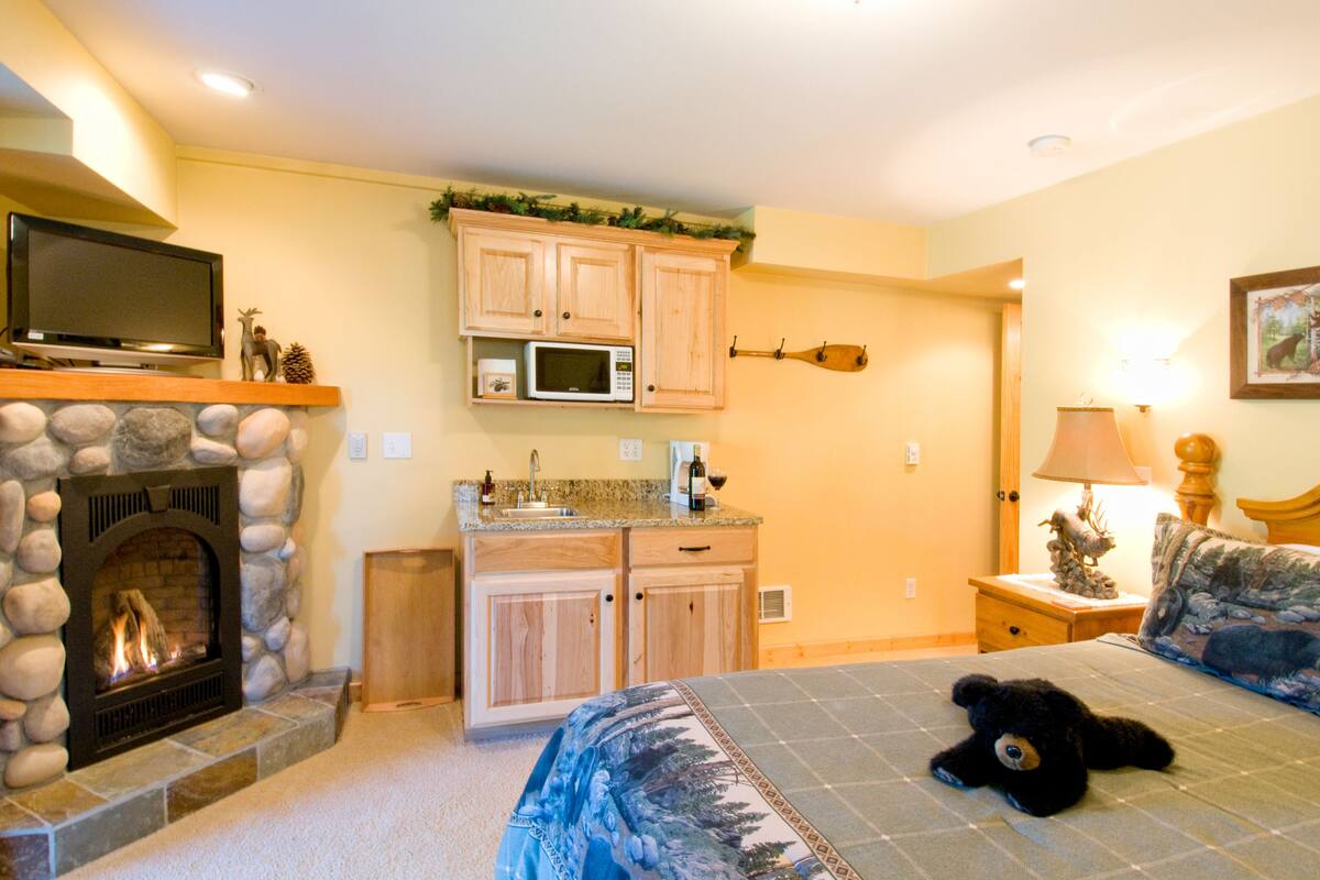 A cozy room with a fireplace, a bed with a quilt, a television, a microwave, and a small kitchen area.