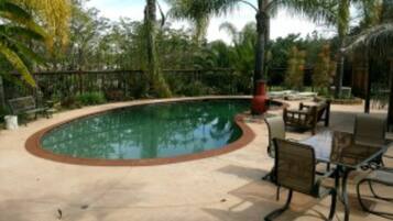 Una piscina curvada con borde rojo, rodeada de palmeras y un patio con mobiliario exterior.