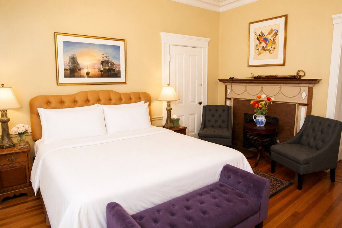 A hotel room with a large bed, two armchairs, a small table with a vase of flowers, and a fireplace.